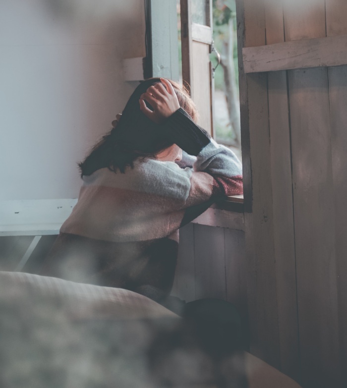 Frau in einer Krise schaut aus dem Fenster. Sie stützt den Kopf mit den Händen ab. Die Farben des Bildes sind eher gehalten bis dunkel.